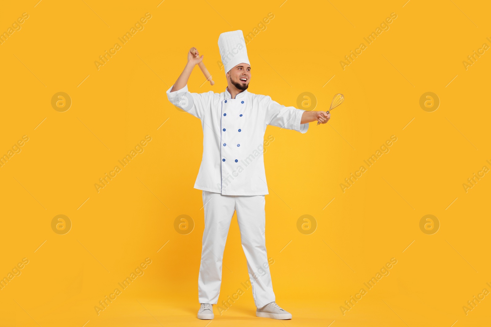 Photo of Happy professional confectioner in uniform holding rolling pin and whisk on yellow background