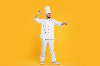 Photo of Happy professional confectioner in uniform holding rolling pin and whisk on yellow background