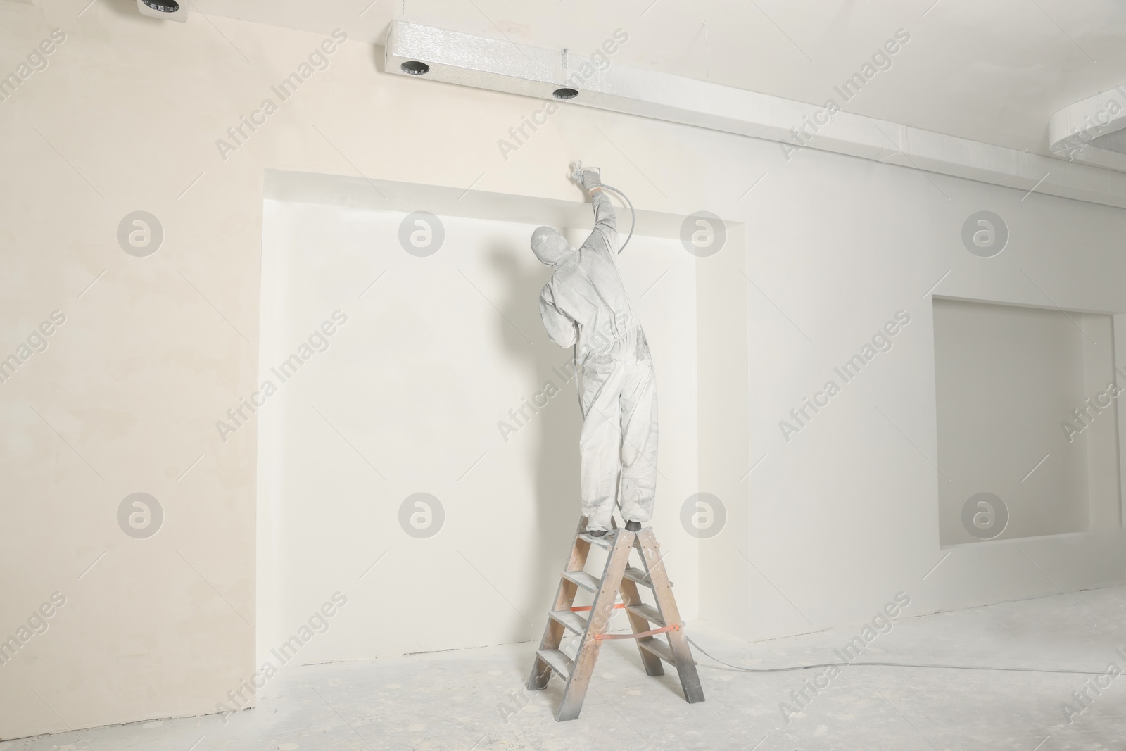 Photo of Decorator in protective overalls painting wall with spray gun indoors