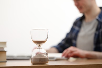 Hourglass with flowing sand on desk. Man taking notes indoors, selective focus