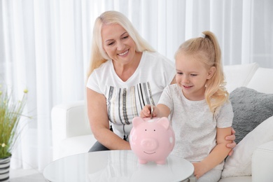 Photo of Little girl putting money into piggy bank and her grandmother at table