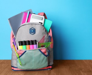 Photo of Backpack with different school stationery on wooden floor near light blue wall, space for text