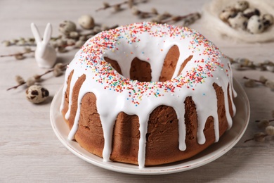 Glazed Easter cake with sprinkles, decorative bunny, quail eggs and willow branches on white wooden table, closeup