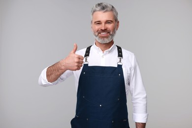 Photo of Happy man in kitchen apron showing thumbs up on grey background. Mockup for design