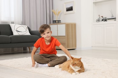 Photo of Little boy petting cute ginger cat on soft carpet at home