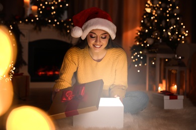 Photo of Young woman wearing Santa hat opening Christmas gift on floor at home