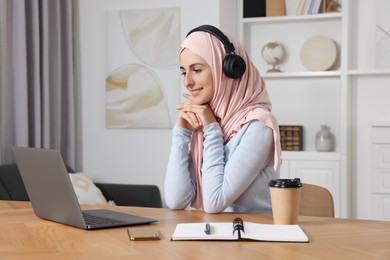 Muslim woman in hijab using laptop at wooden table in room