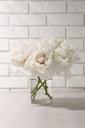 Beautiful peonies in glass vase on white table near brick wall