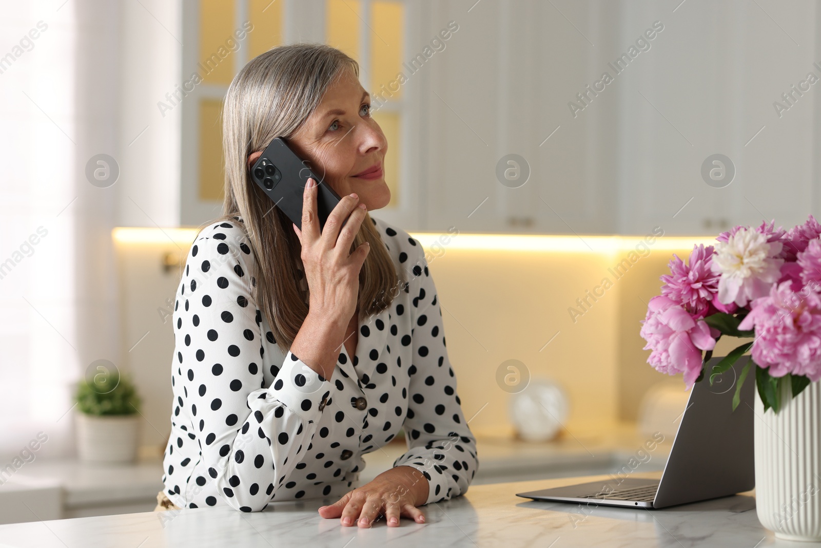 Photo of Senior woman talking on phone at white table indoors