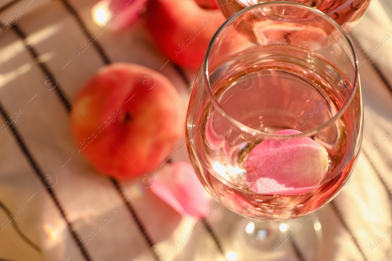 Photo of Glass of delicious rose wine with petals and peaches on white picnic blanket, closeup. Space for text