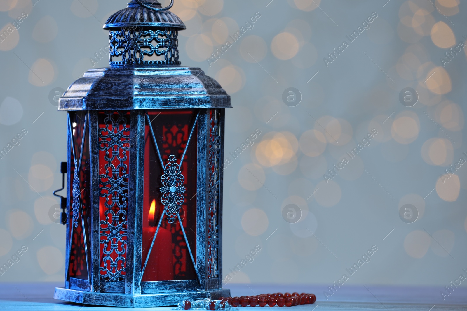 Photo of Arabic lantern and misbaha on table against blurred lights, space for text