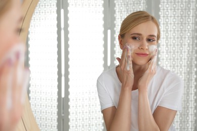 Photo of Happy young woman applying cleansing foam onto face near mirror in bathroom