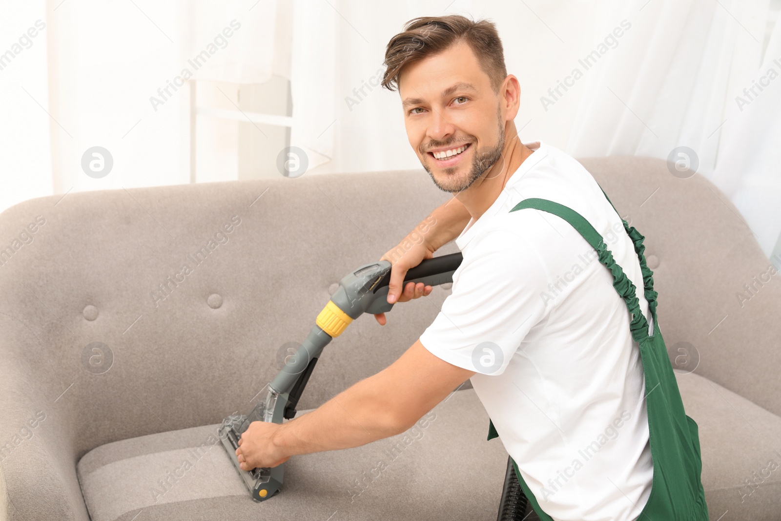 Photo of Male janitor removing dirt from sofa with upholstery cleaner indoors