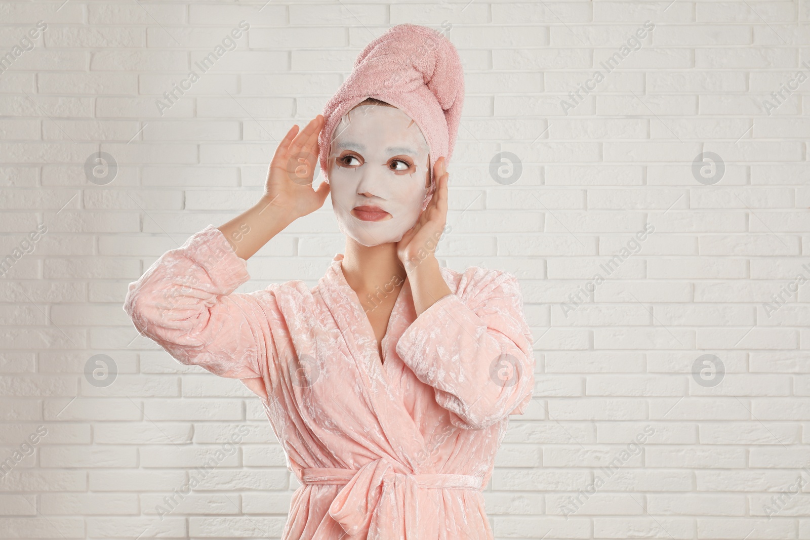 Photo of Young woman in bathrobe with cotton facial mask near white brick wall