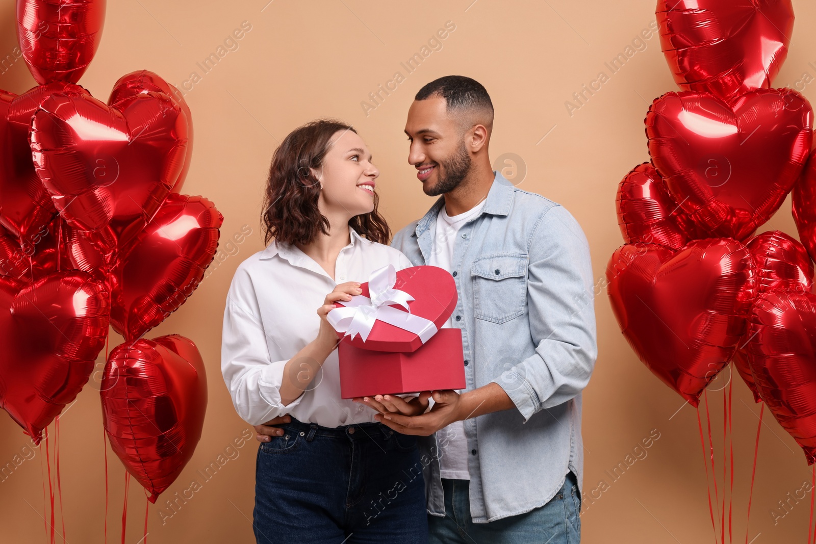 Photo of Lovely couple with gift box near heart shaped air balloons on beige background. Valentine's day celebration