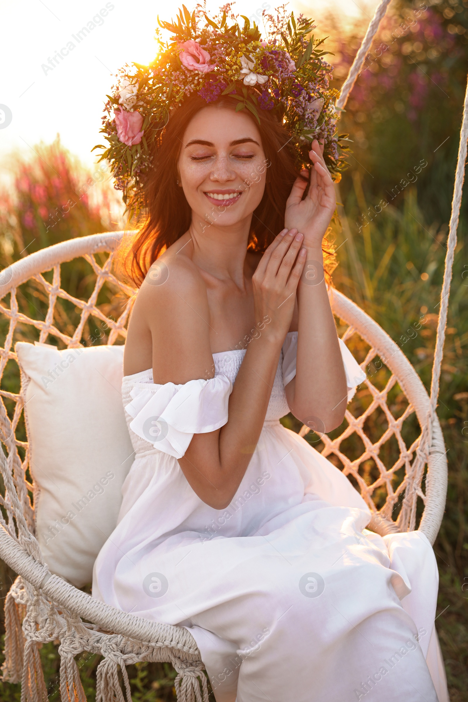 Photo of Young woman wearing wreath made of beautiful flowers on swing chair outdoors at sunset