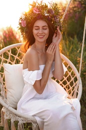 Young woman wearing wreath made of beautiful flowers on swing chair outdoors at sunset