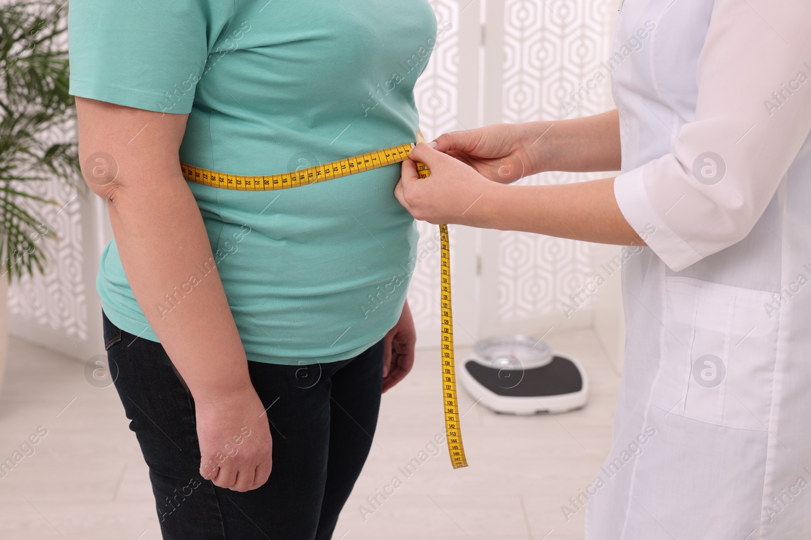 Photo of Nutritionist measuring overweight woman's waist with tape in clinic, closeup