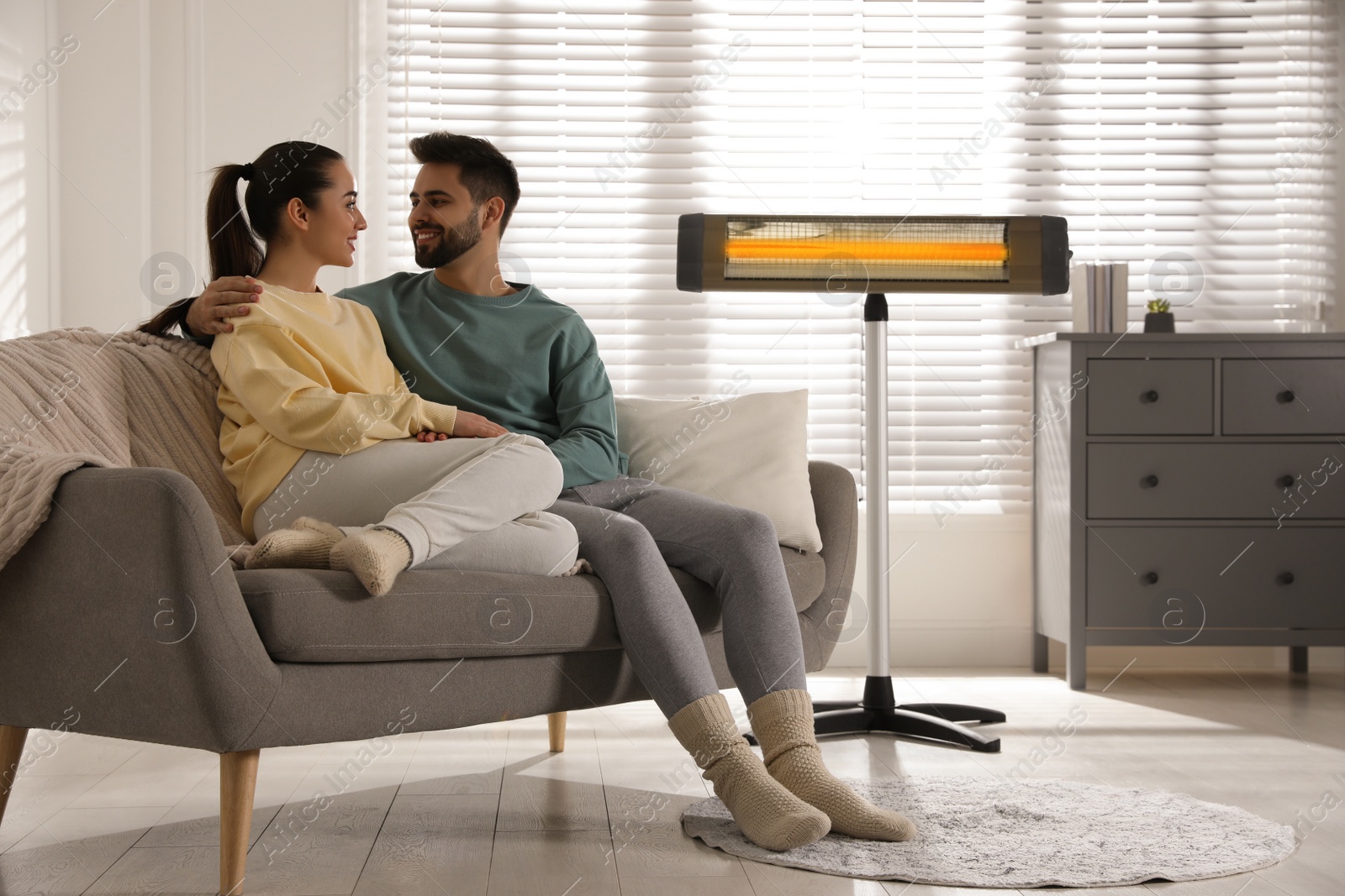 Photo of Young couple sitting on sofa near electric heater at home