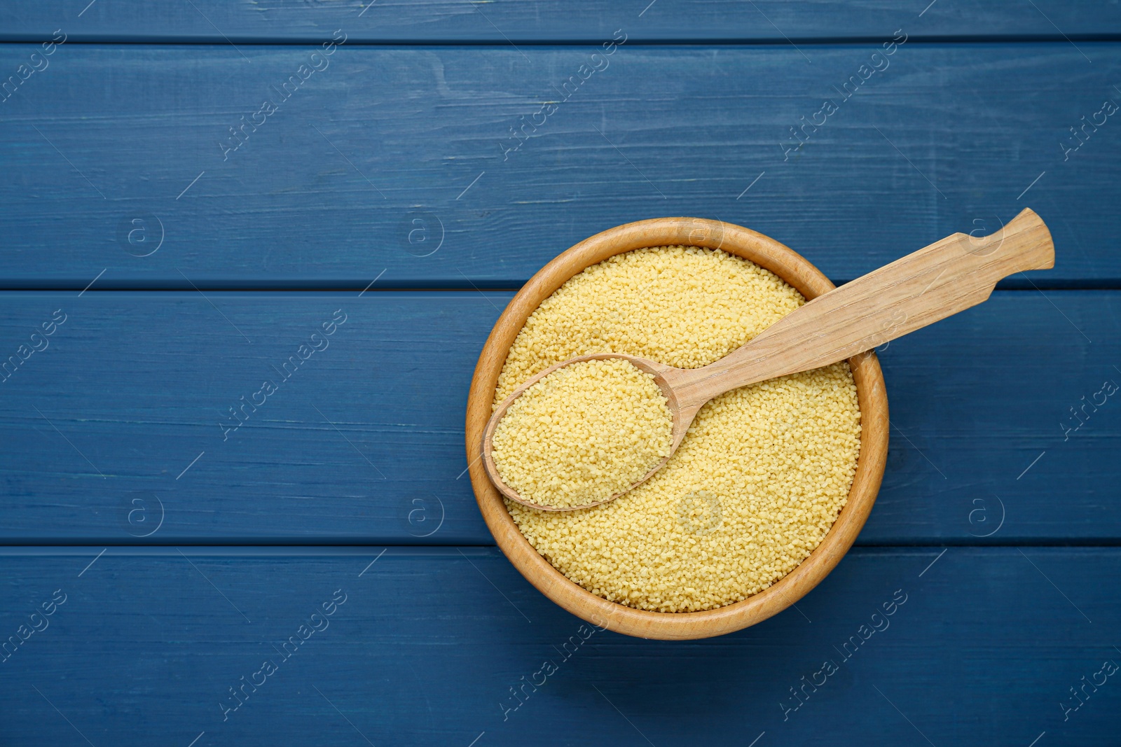 Photo of Bowl and spoon with raw couscous on blue wooden table, top view. Space for text