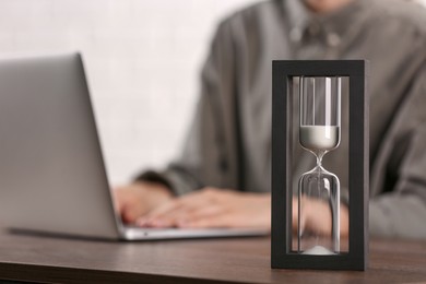 Photo of Hourglass with flowing sand on wooden table, selective focus. Man using laptop indoors