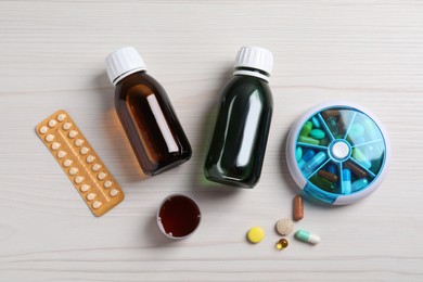 Photo of Bottles of syrup, measuring cup and pills on white wooden table, flat lay. Cold medicine