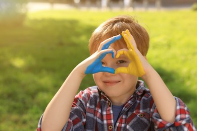Photo of Little boy making heart with his hands painted in Ukrainian flag colors outdoors, space for text. Love Ukraine concept
