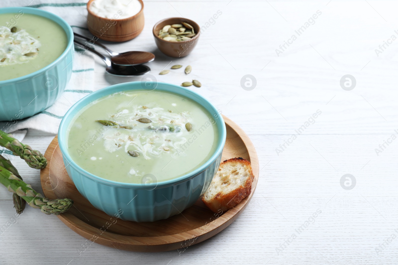 Photo of Bowl of delicious asparagus soup served on white wooden table, space for text