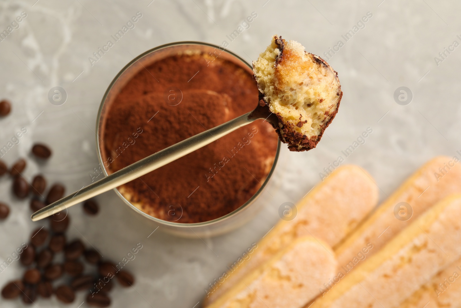 Photo of Tasty tiramisu in glass, coffee beans, spoon and biscuits on light grey table, flat lay