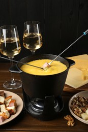 Fondue pot with melted cheese, glasses of wine, forks and different products on wooden table