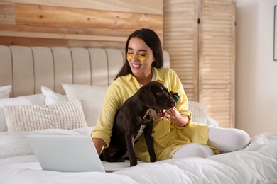 Photo of Young woman playing with her dog while working on laptop in bedroom. Home office concept