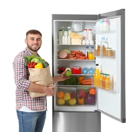 Young man with bag of groceries and apple near open refrigerator on white background