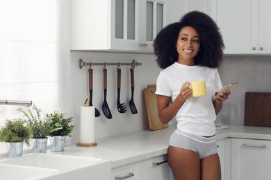 Beautiful woman in stylish underwear holding smartphone and cup of drink indoors