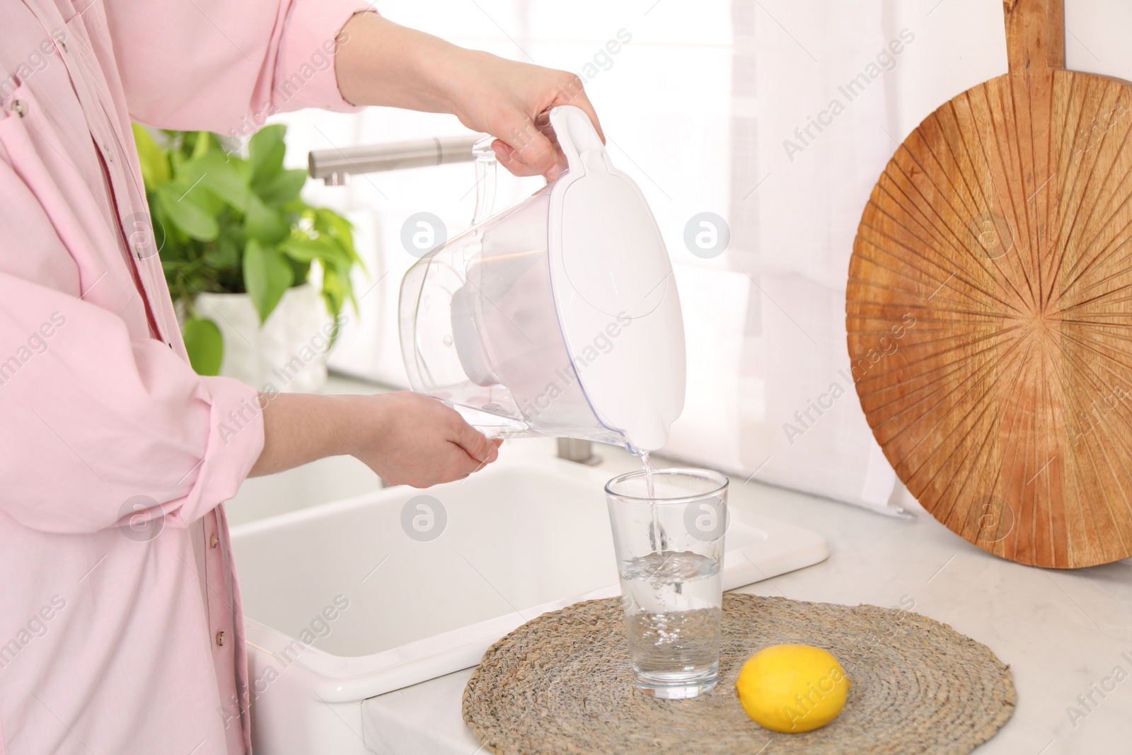 Photo of Woman pouring water from filter jug into glass in kitchen, closeup