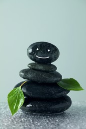 Photo of Stack of stones with drawn happy face, green leaves and water drops on table against grey background. Zen concept