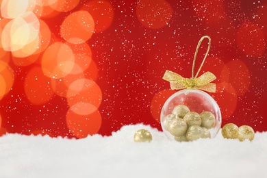 Image of Beautiful transparent Christmas ornament with small golden balls on snow against red background, bokeh effect. Space for text