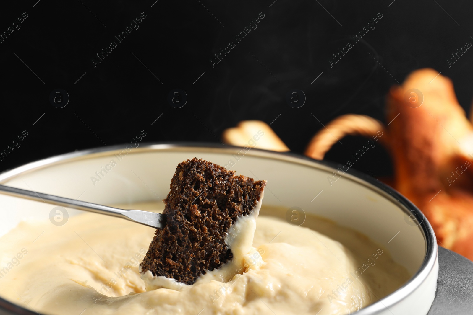 Photo of Dipping piece of bread into fondue pot with melted cheese on black background, closeup