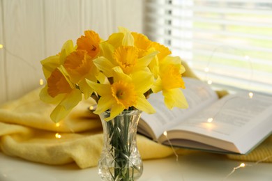 Photo of Beautiful yellow daffodils in vase, book and festive lights on windowsill