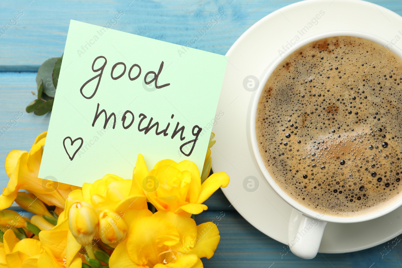 Photo of Cup of aromatic coffee, beautiful yellow freesias and Good Morning note on light blue wooden table, flat lay