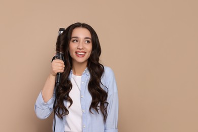 Photo of Happy woman using curling hair iron on beige background. Space for text