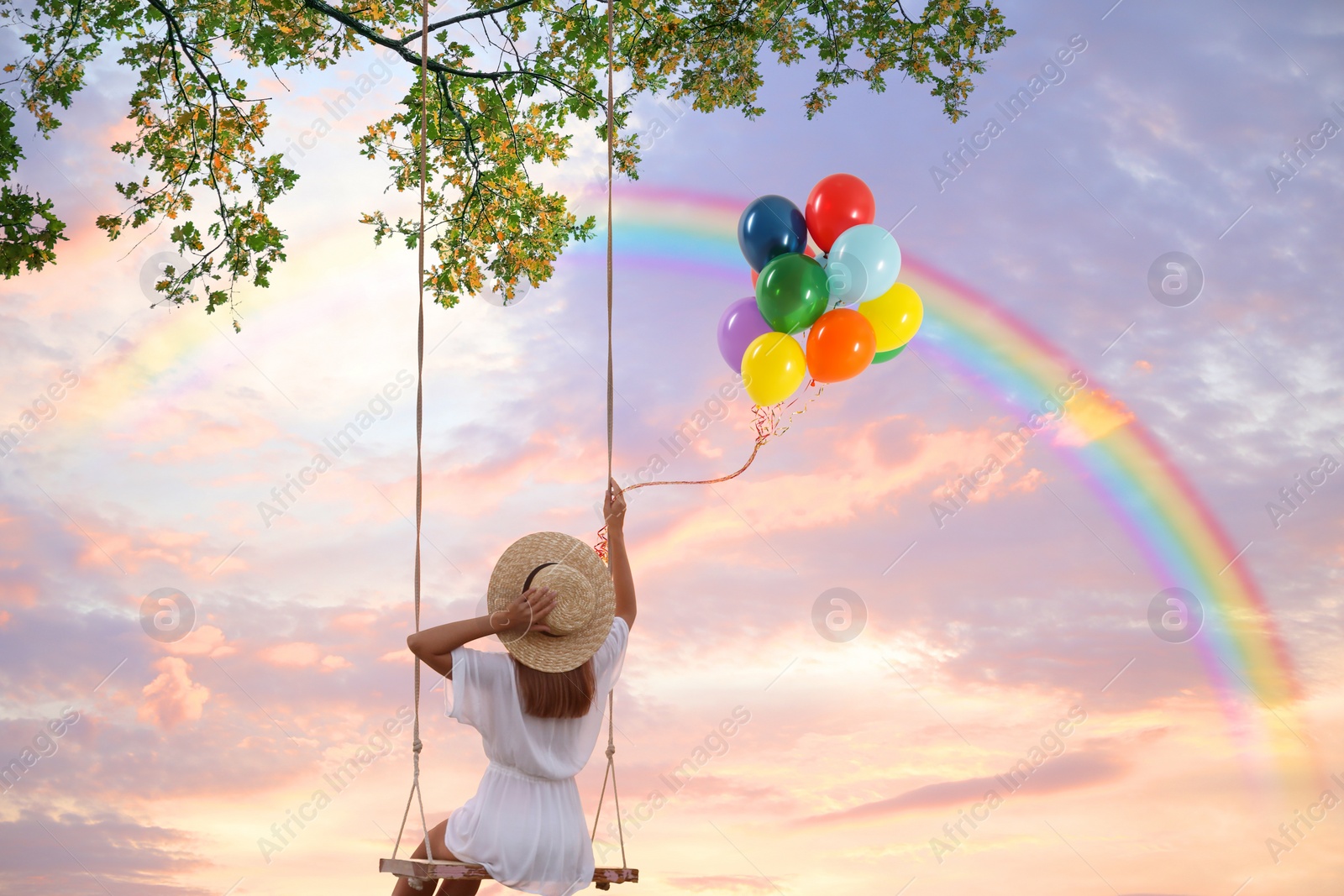 Image of Dream world. Young woman with bright balloons swinging, rainbow in sunset sky on background