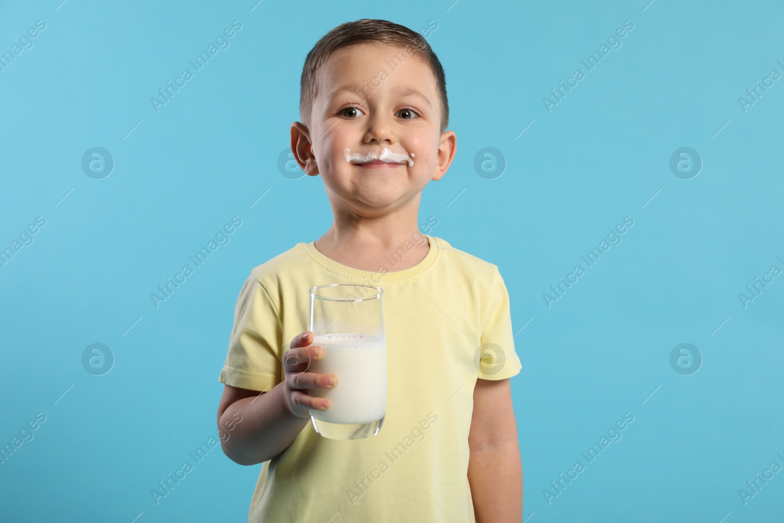 Photo of Cute boy with milk mustache holding glass of tasty dairy drink on light blue background