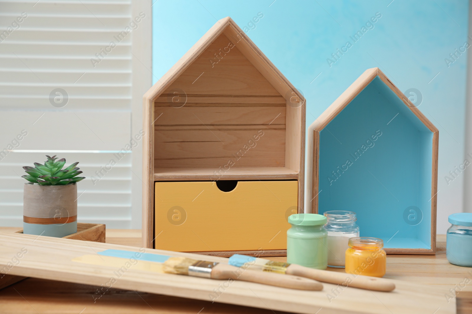 Photo of House shaped shelves, brushes and jars of paints on table indoors