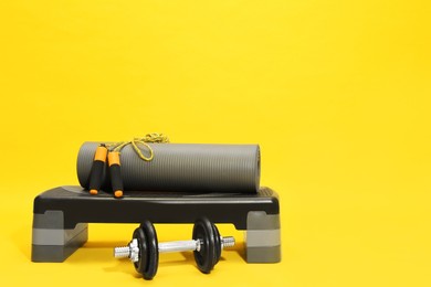 Photo of Step platform, dumbbell, mat and jump rope on yellow background, space for text. Sports equipment