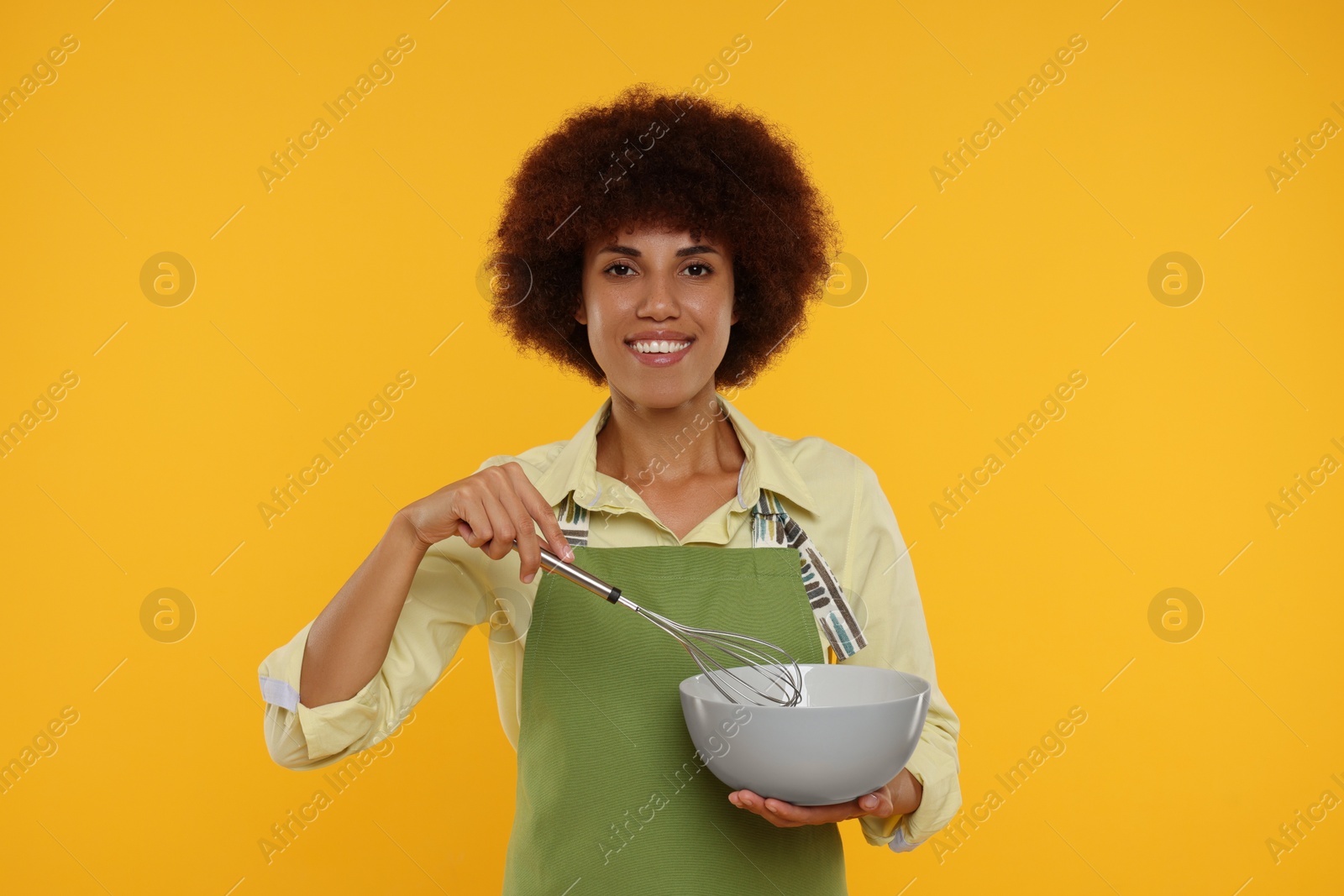 Photo of Happy young woman in apron holding bowl and whisk on orange background