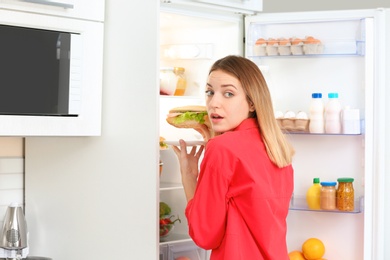 Photo of Emotional young woman taking sandwich from refrigerator in kitchen. Failed diet