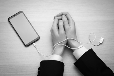 Image of Top view of woman with her hands tangled in charging cable of smartphone at wooden table, black and white effect. Internet addiction