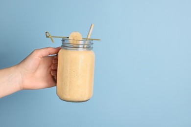 Photo of Woman holding mason jar with tasty smoothie, banana and straw on light blue background, closeup. Space for text