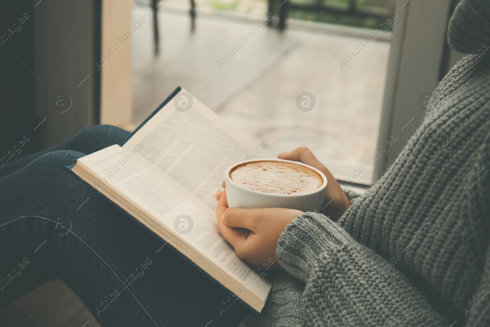 Image of Woman with cup of coffee reading book near window indoors, closeup