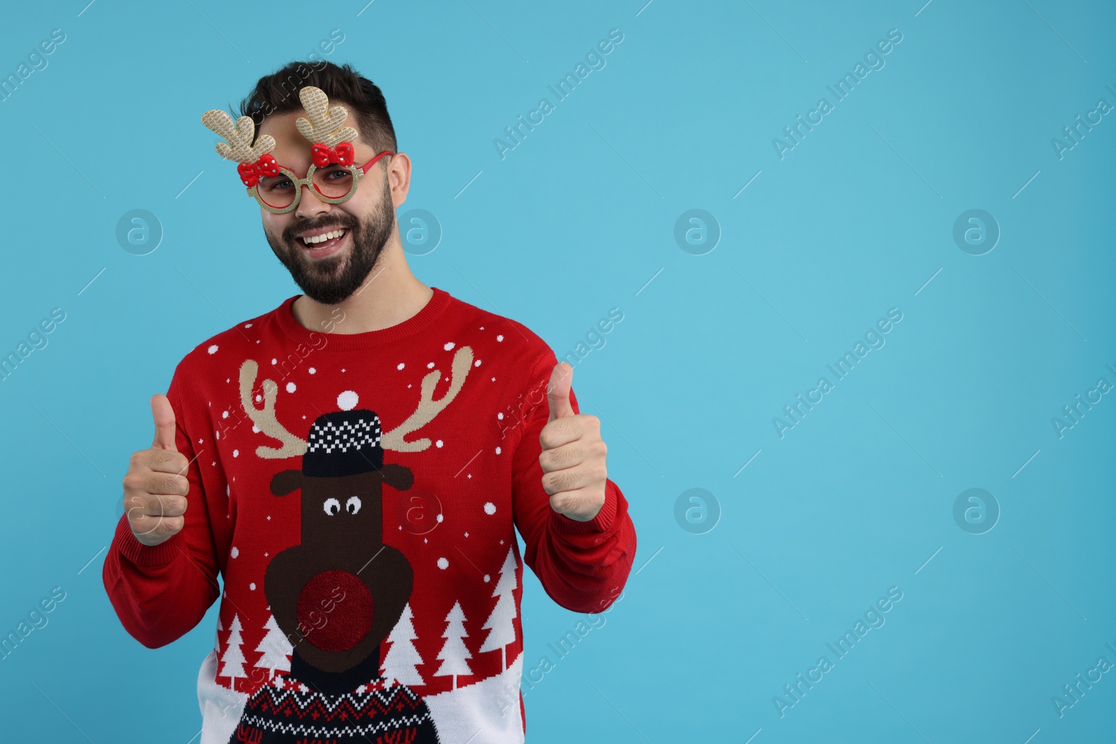 Photo of Happy young man in Christmas sweater and funny glasses showing thumbs up on light blue background. Space for text